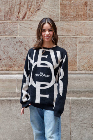 Woman wearing a black sweater with beige 'New York' logo against a stone wall.