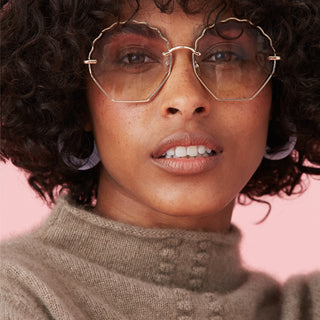 Close-up portrait of a woman with curly hair wearing stylish sunglasses and a cozy sweater against a pink background.