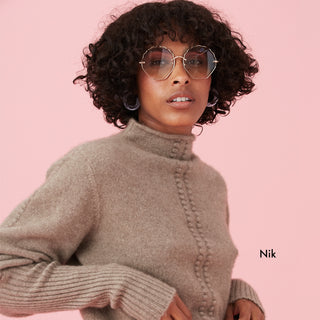 Person in a light brown sweater with curly hair and stylish glasses, standing against a pink background