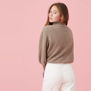A young woman with long hair wearing a brown sweater and white pants, looking back over her shoulder against a pink background.