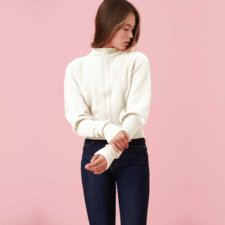 A model wearing a cream-colored sweater and dark jeans against a pink background.