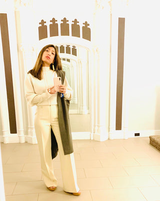 A person stands in front of a mirror, wearing a cream-colored outfit with a dark scarf, taking a selfie.